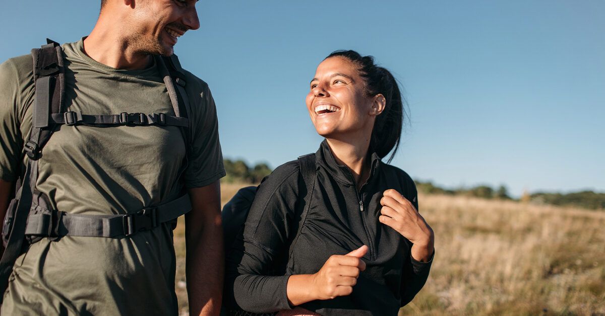 A man and woman wearing weighted backpacks walking through a sunlit field.