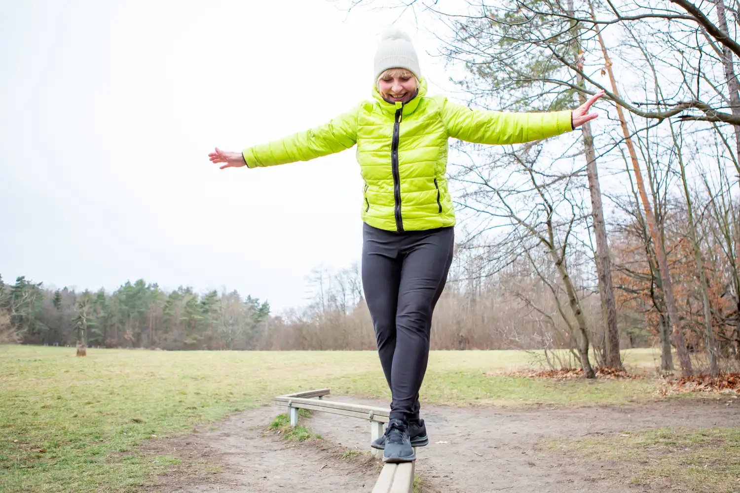 An older woman balancing on a wooden beam in a park with her arms outstretched.