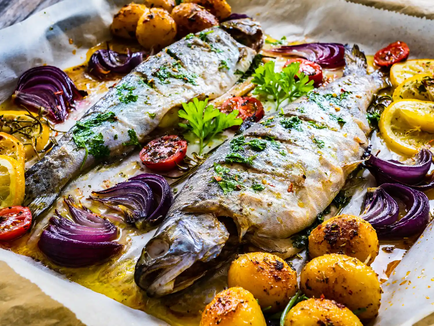 Whole baked fish with roasted potatoes, cherry tomatoes, and lemon slices on parchment paper.