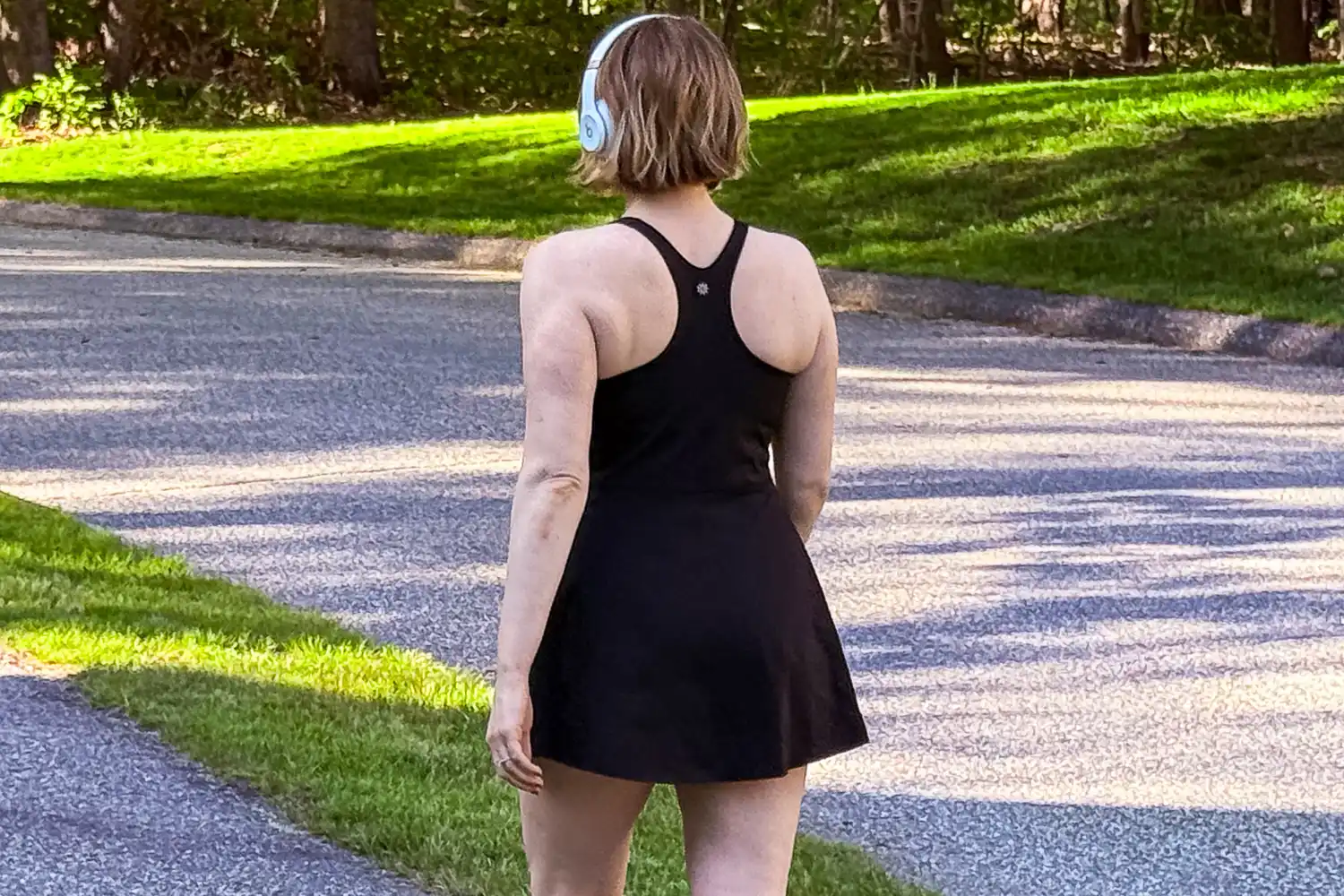 A woman in a black athletic dress and headphones walking in a sunny park.