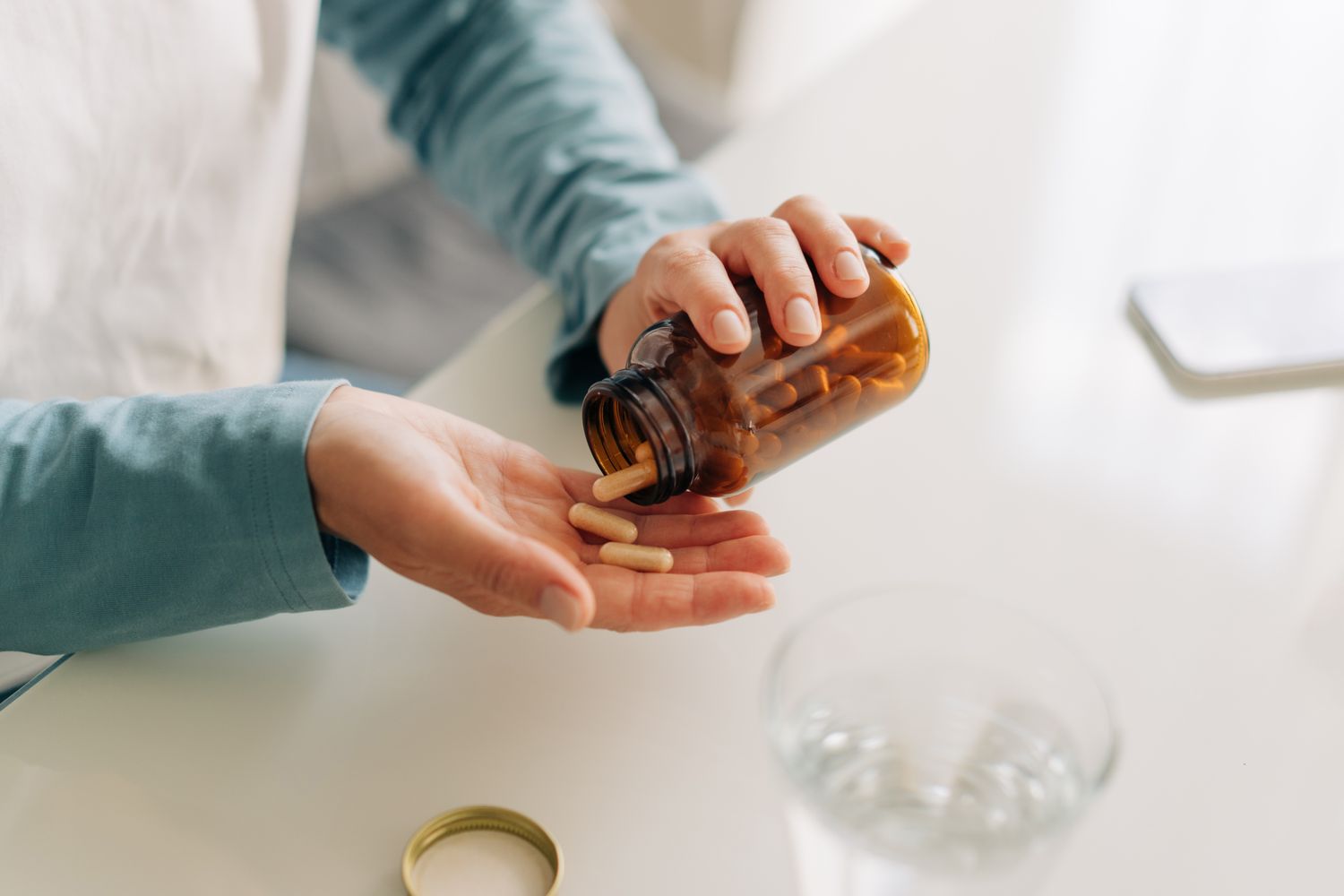 A person pouring capsules from a brown bottle into their palm on a white table near a glass of water.