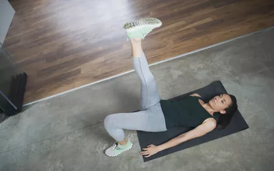 A woman performing a single-leg glute bridge on a black yoga mat in a home setting.