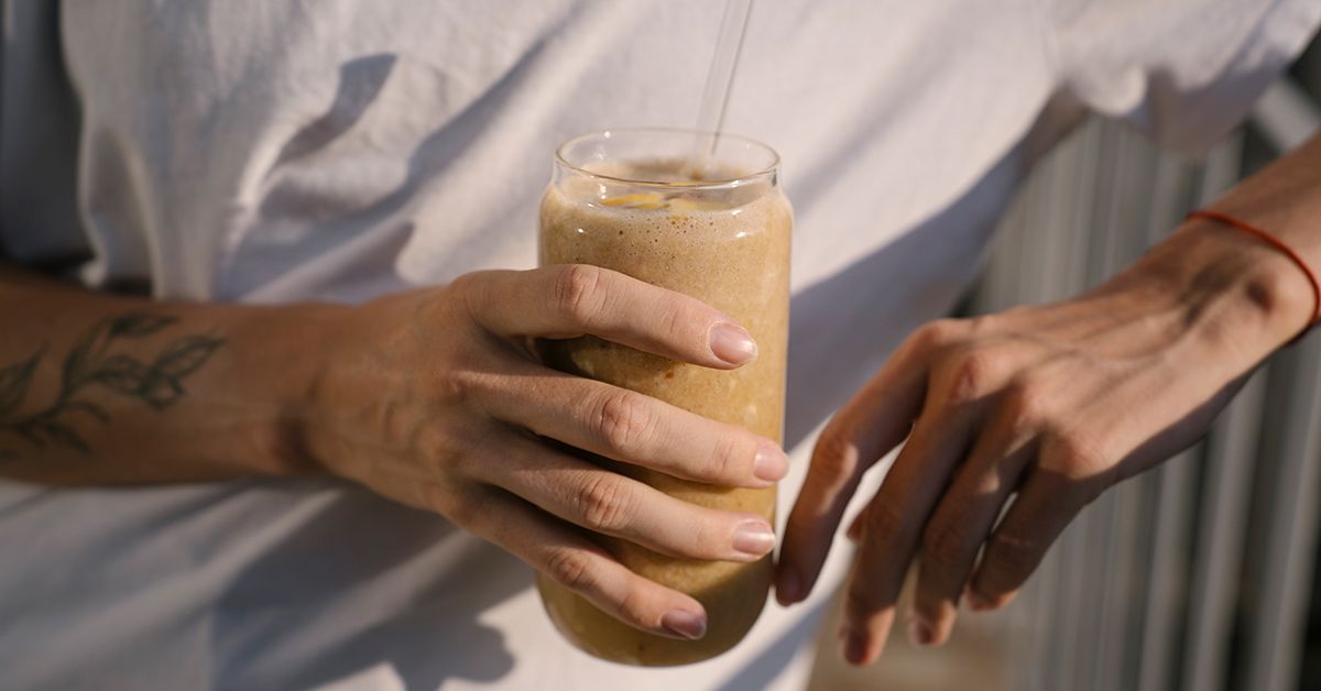 A person holding a glass of a creamy, blended smoothie in a sunlit setting.