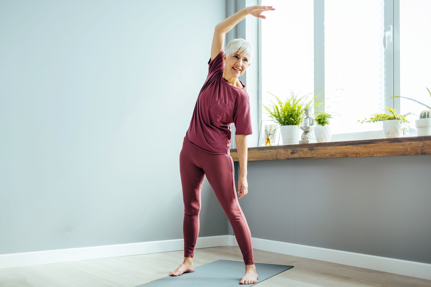 An older woman with white hair stretching on a yoga mat in a sunlit room filled with plants.