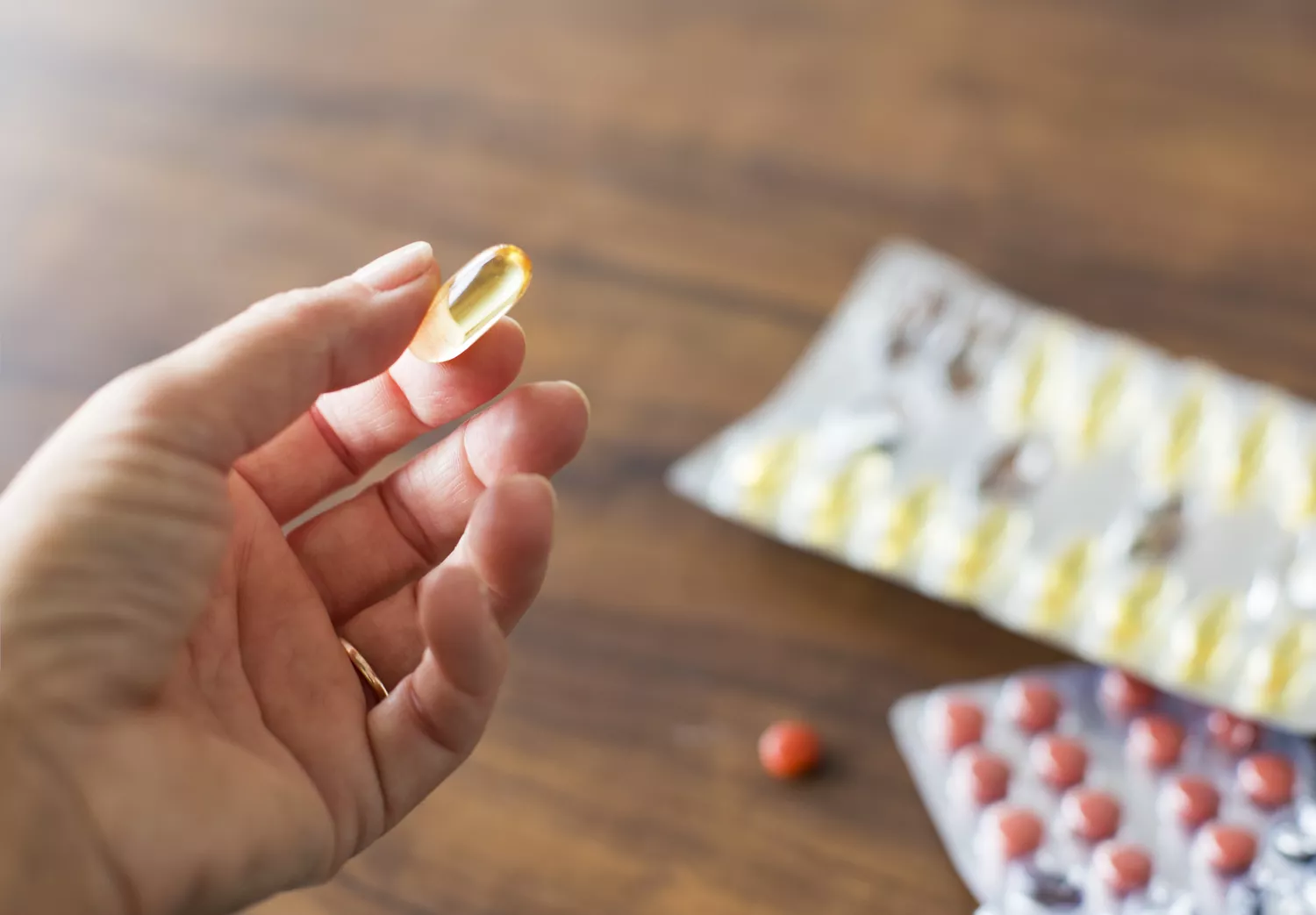 A close-up of a person holding a yellow omega-3 softgel capsule with blurred supplement packaging in the background.