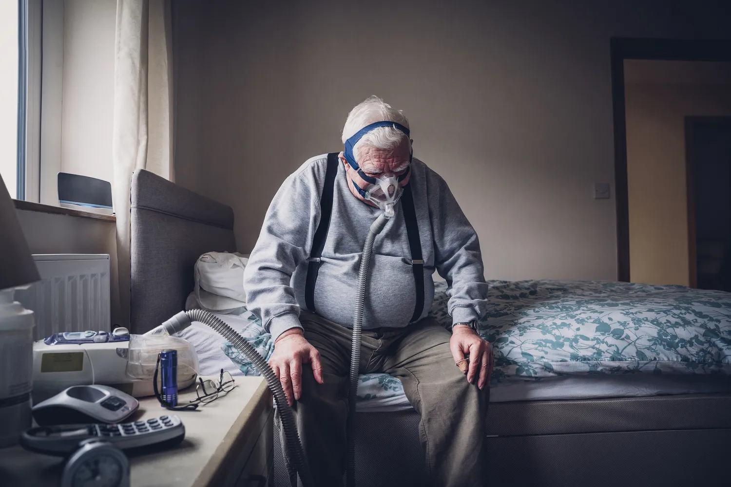 A senior man using a CPAP machine with tubing in a dimly lit bedroom setting to illustrate nighttime treatment.