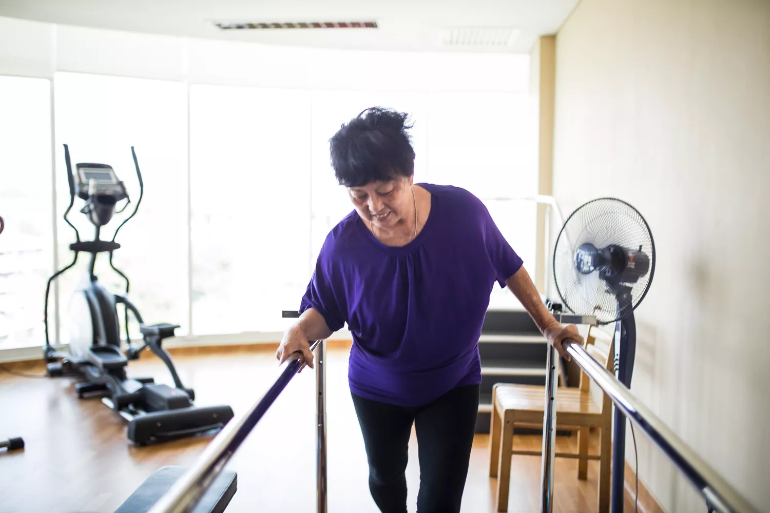 An elderly woman using parallel bars for assisted walking in a bright physical therapy gym.