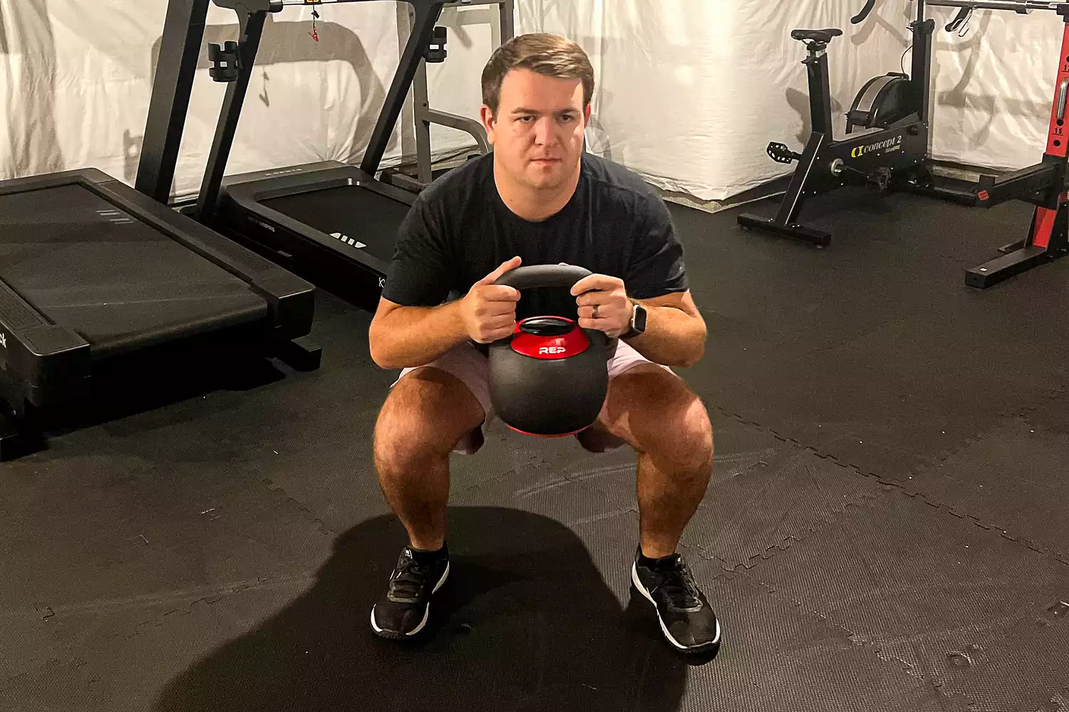 A man performing a goblet squat using an adjustable kettlebell in a home gym setting.
