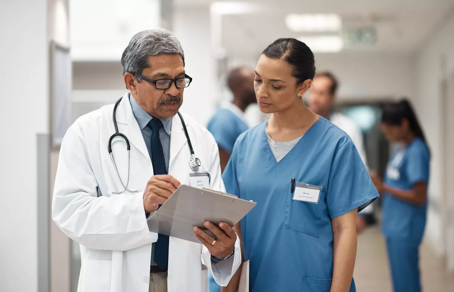 Two healthcare providers in white coats and scrubs consulting each other in a bright hospital hallway.