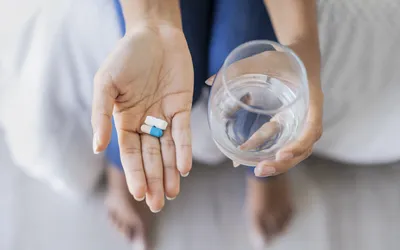 A person holding two supplement capsules in one hand and a glass of water in the other.