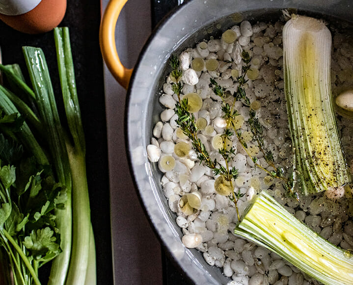A person preparing a bowl of soaked beans with fresh herbs on a wooden kitchen counter.