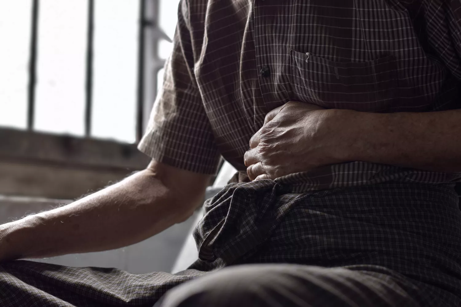 An elderly man in a plaid shirt sits with his hand pressed to his abdomen, suggesting severe medical discomfort.
