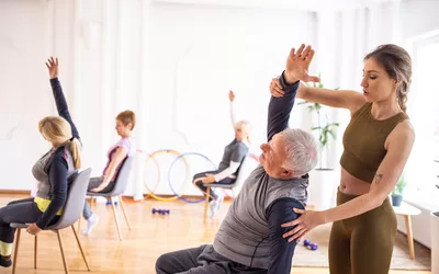 A female fitness instructor assisting an older man with seated arm stretches in a group class.