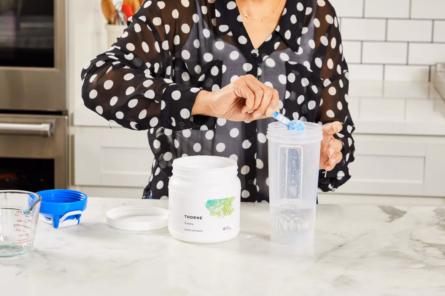 A woman in a kitchen adding a scoop of white creatine powder into a shaker bottle.