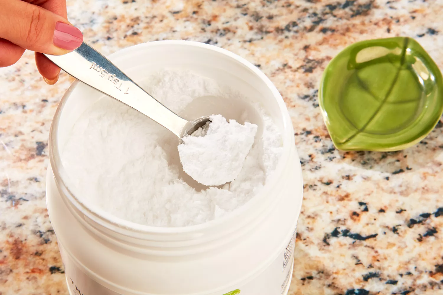 A hand using a measuring spoon to scoop fine white creatine powder from a jar.