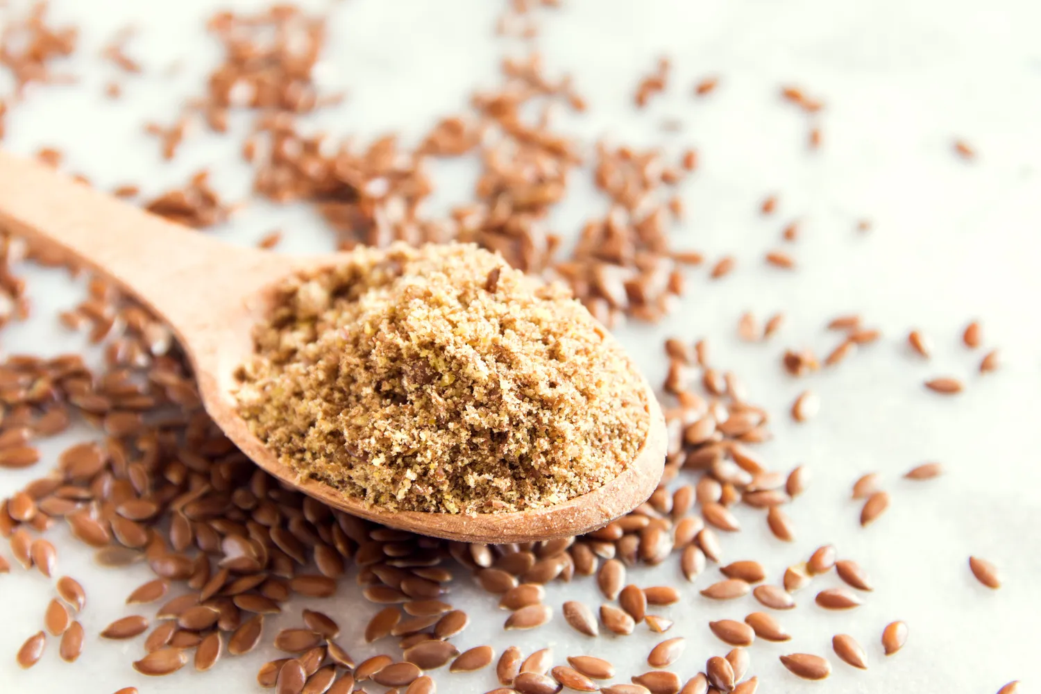 Ground flaxseed on a wooden spoon surrounded by whole flaxseeds.